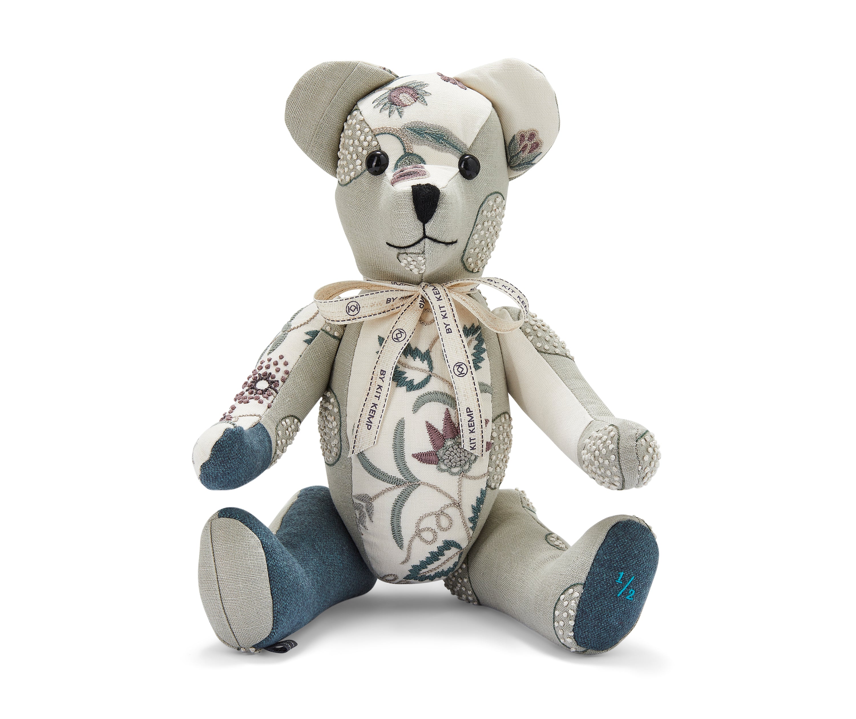 The image shows a handmade teddy bear toy. The bear is primarily made from patterned fabric featuring floral and leaf designs in shades of blue, green, and maroon. It has a light-coloured face with embroidered features. The bear is wearing a decorative ribbon tied in a bow around its neck. The bear's limbs are posed in a sitting position.