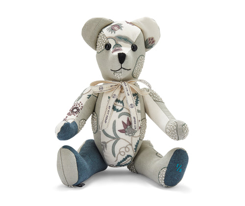 The image shows a handmade teddy bear toy. The bear is primarily made from patterned fabric featuring floral and leaf designs in shades of blue, green, and maroon. It has a light-coloured face with embroidered features. The bear is wearing a decorative ribbon tied in a bow around its neck. The bear's limbs are posed in a sitting position.