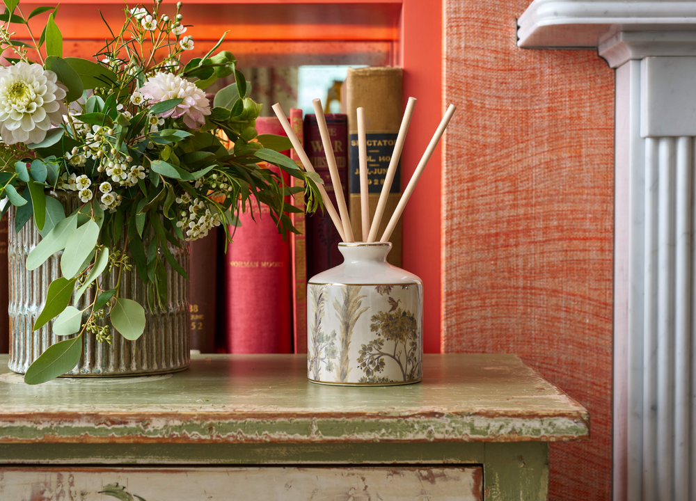 A ceramic diffuser with a tall trees design next to a vase of flowers on a wooden table