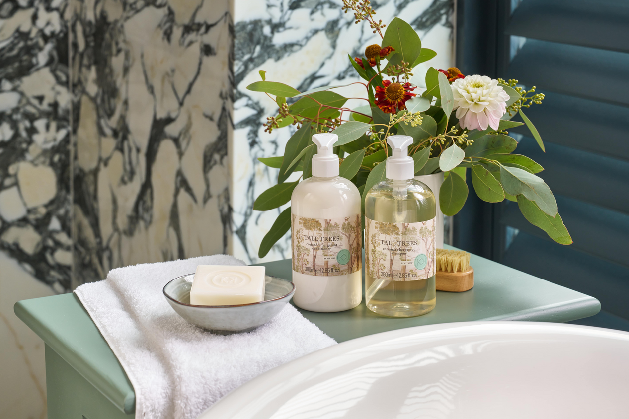 Life style image of Tall trees shampoo and conditioner, presented beside roll top bath, on a sage green table.