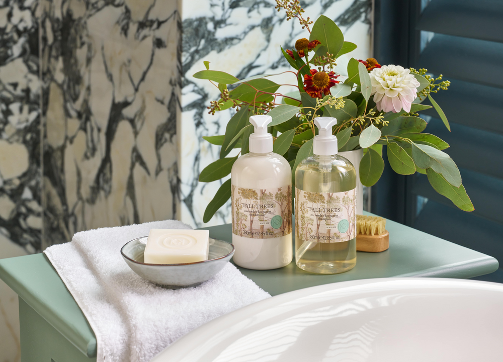 Life style image of Tall trees shampoo and conditioner, presented beside roll top bath, on a sage green table.