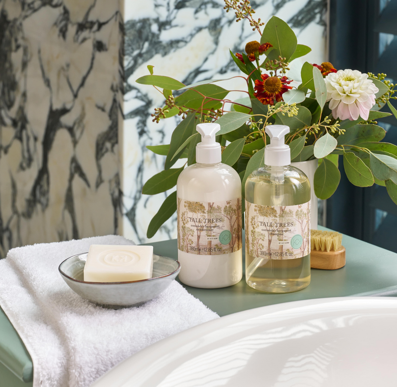 Life style image of Tall trees shampoo and conditioner, presented beside roll top bath, on a sage green table.