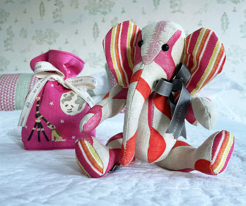 Elephant patchwork toy made out of hot pink, and orange rik rak fabric , on a natural background, with hot pink lavender bag, embroidered with moon dog design, (delivered as a gift, in bright pink, and yellow gift box). Presented on a bed with a white bedspread, and a pink and green cushion in the back ground.