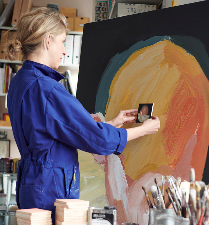 Person in a blue jumpsuit painting on an easel with a palette and brushes in the foreground. Holly Frean