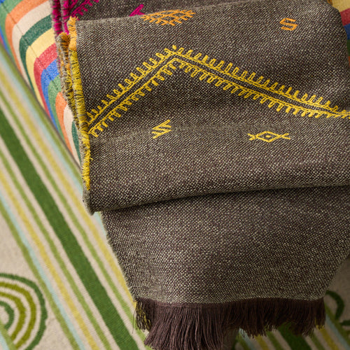 A folded dark olive woven throw with fringe and bright yellow and magenta geometric embroidery