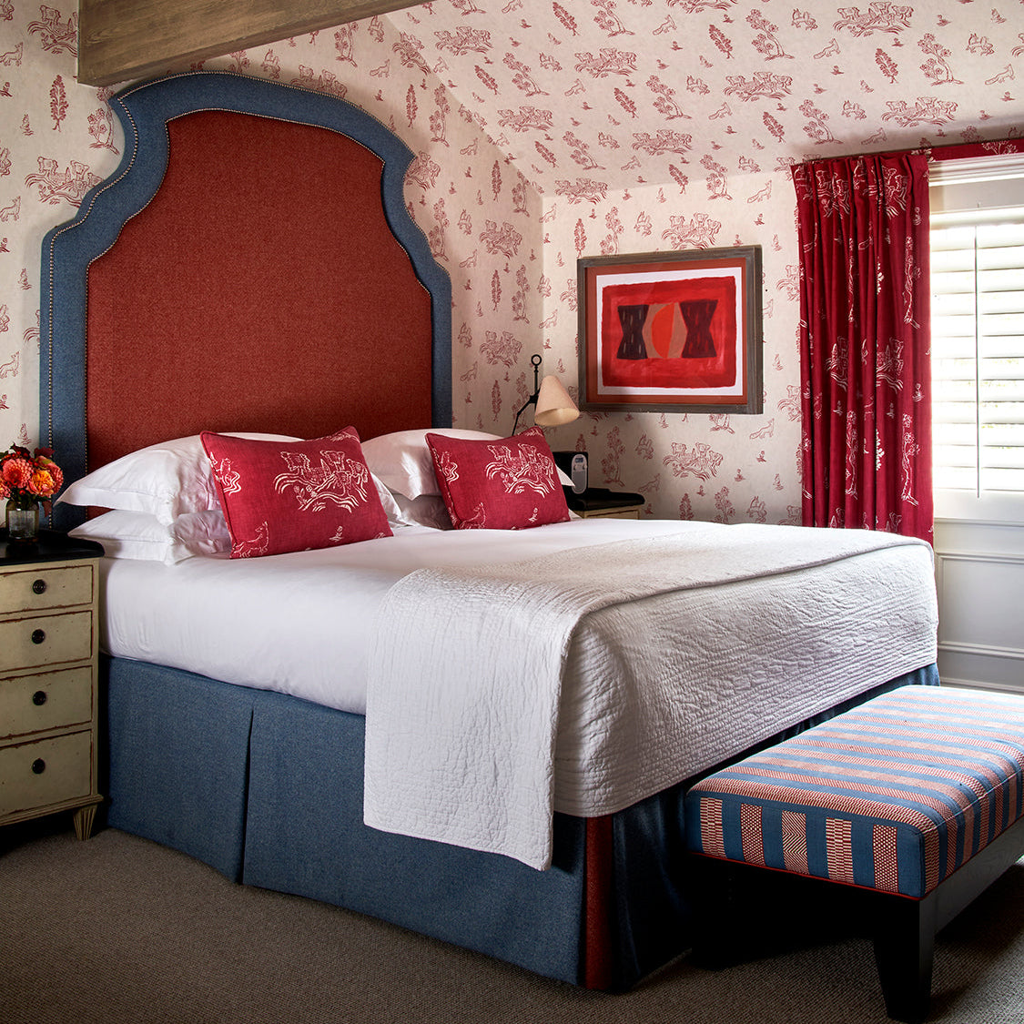 Number 16 hotel bedroom featuring Friendly Folk theme wallpaper and cushions, with Ellie Headboard - Two Colour Fabric in red and blue, completed with End Of Bed Stool. Designed by Kit Kemp Design Studio.