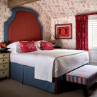 Number 16 hotel bedroom featuring Friendly Folk theme wallpaper and cushions, with Ellie Headboard - Two Colour Fabric in red and blue, completed with End Of Bed Stool. Designed by Kit Kemp Design Studio.