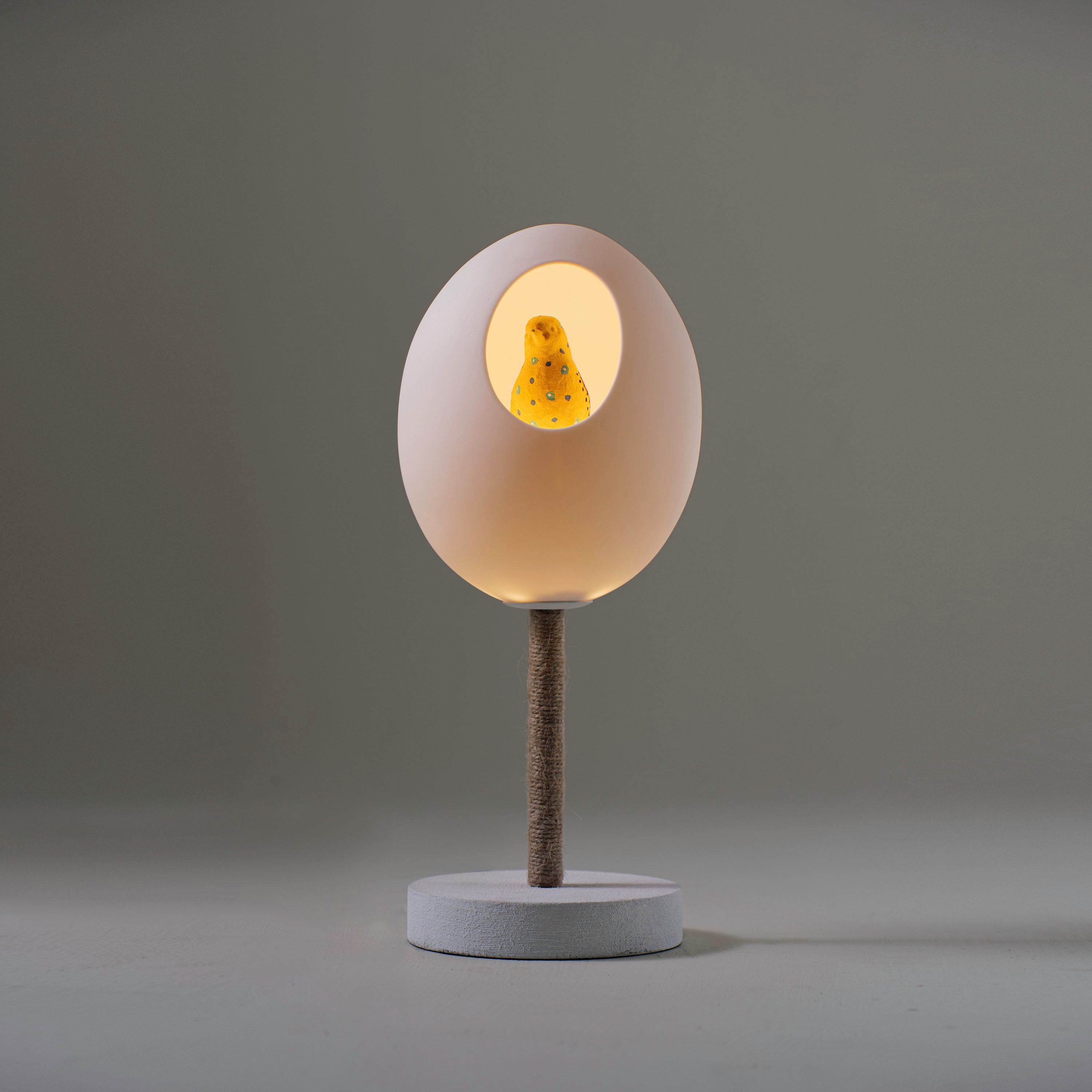 A table lamp in the shape of a large white egg with a yellow bird in side.  The lamp sitting on a white base