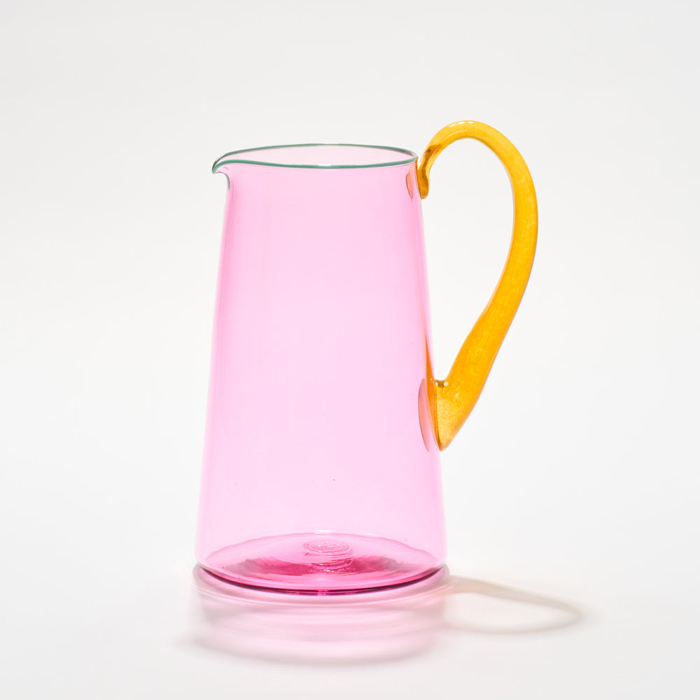A vibrant pink hand-blown glass jug with a bold transparent pink body, a teal lip, and an orange handle, each is a handcrafted piece of art