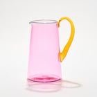 A vibrant pink hand-blown glass jug with a bold transparent pink body, a teal lip, and an orange handle, each is a handcrafted piece of art