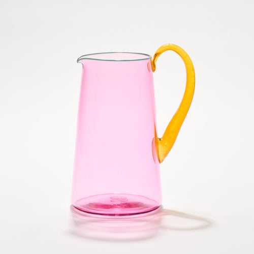 A vibrant pink hand-blown glass jug with a bold transparent pink body, a teal lip, and an orange handle, each is a handcrafted piece of art
