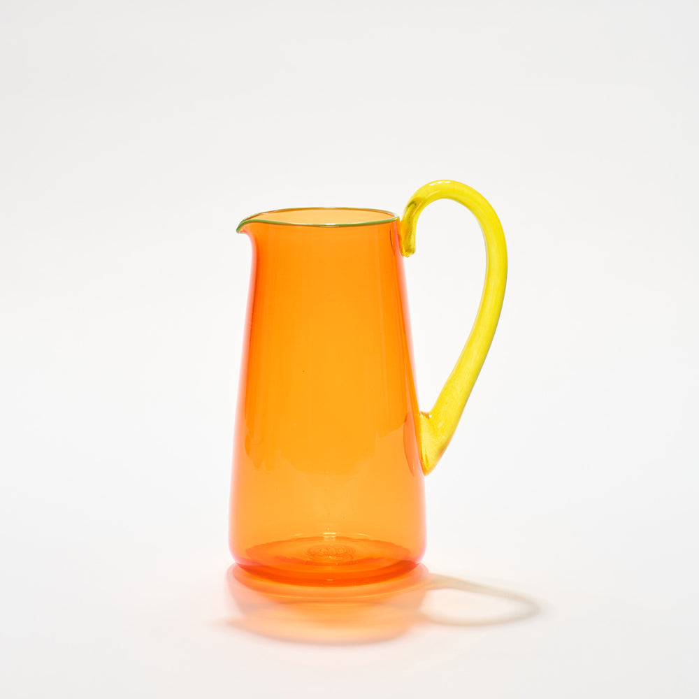 A vibrant hand-blown glass jug with a transparent orange body, green lip, and yellow handle, hand-blown glass made in the UK.