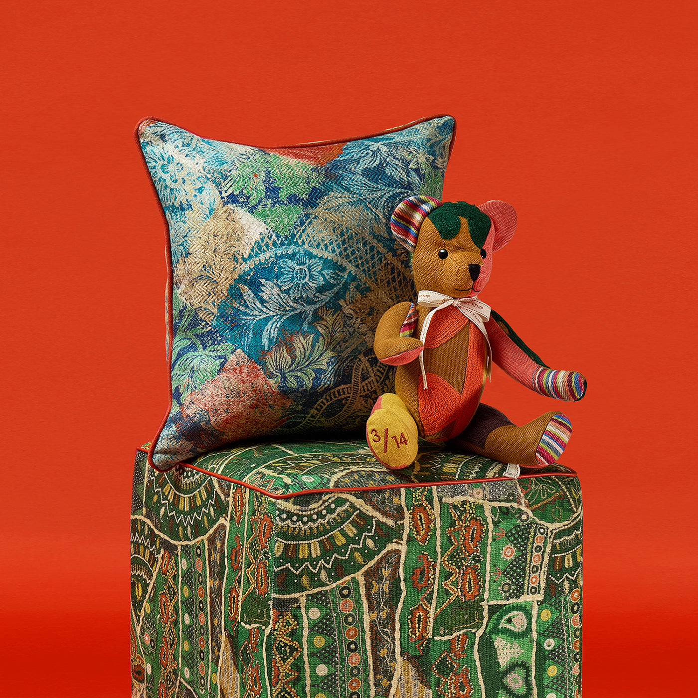 A handmade Large Willow Bear from Kit Kemp’s Patchwork Collection sits on a green patterned Front Row Stool. The bear features a rich ochre body with vibrant patchwork accents in emerald green and coral pink, showcasing premium textile offcuts and a refined, artisanal quality.