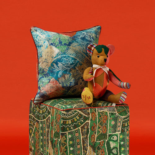 A handmade Large Willow Bear from Kit Kemp’s Patchwork Collection sits on a green patterned Front Row Stool. The bear features a rich ochre body with vibrant patchwork accents in emerald green and coral pink, showcasing premium textile offcuts and a refined, artisanal quality.