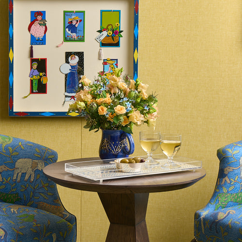 Round table with a vase of flowers, two glasses of white wine, and a tray with fruit in a room with yellow walls and colorful wall art.
