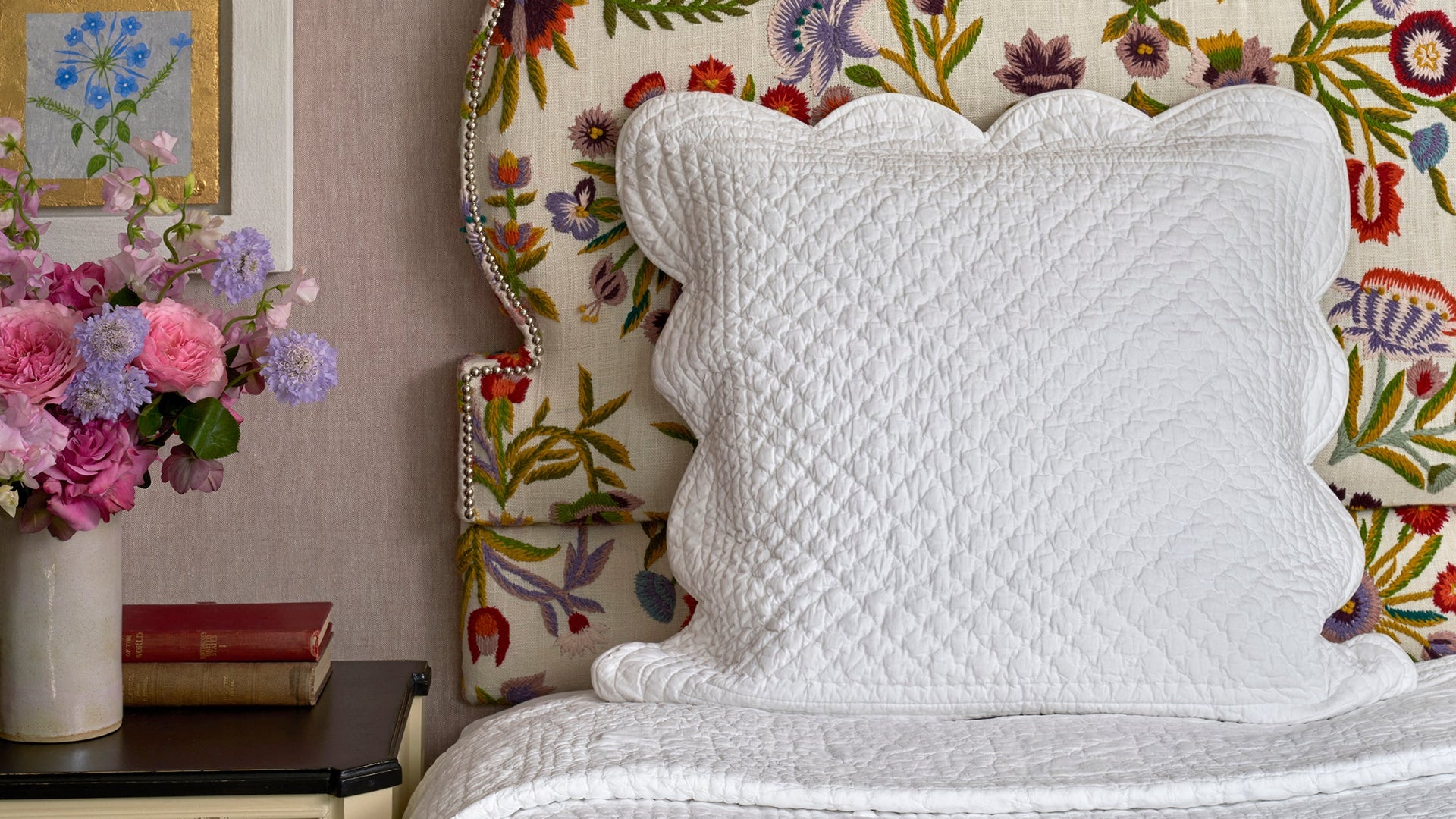 A close-up shot of a bed featuring the Powershake bedding collection by Shop Kit Kemp. The quilted, white cotton pillow has scalloped edges and a border of crisp, concentric channels of stitching. The comforting, cottony pillow provides hotel standard sleep quality and rests against an upholstered headboard, in bespoke Kit Kemp design.