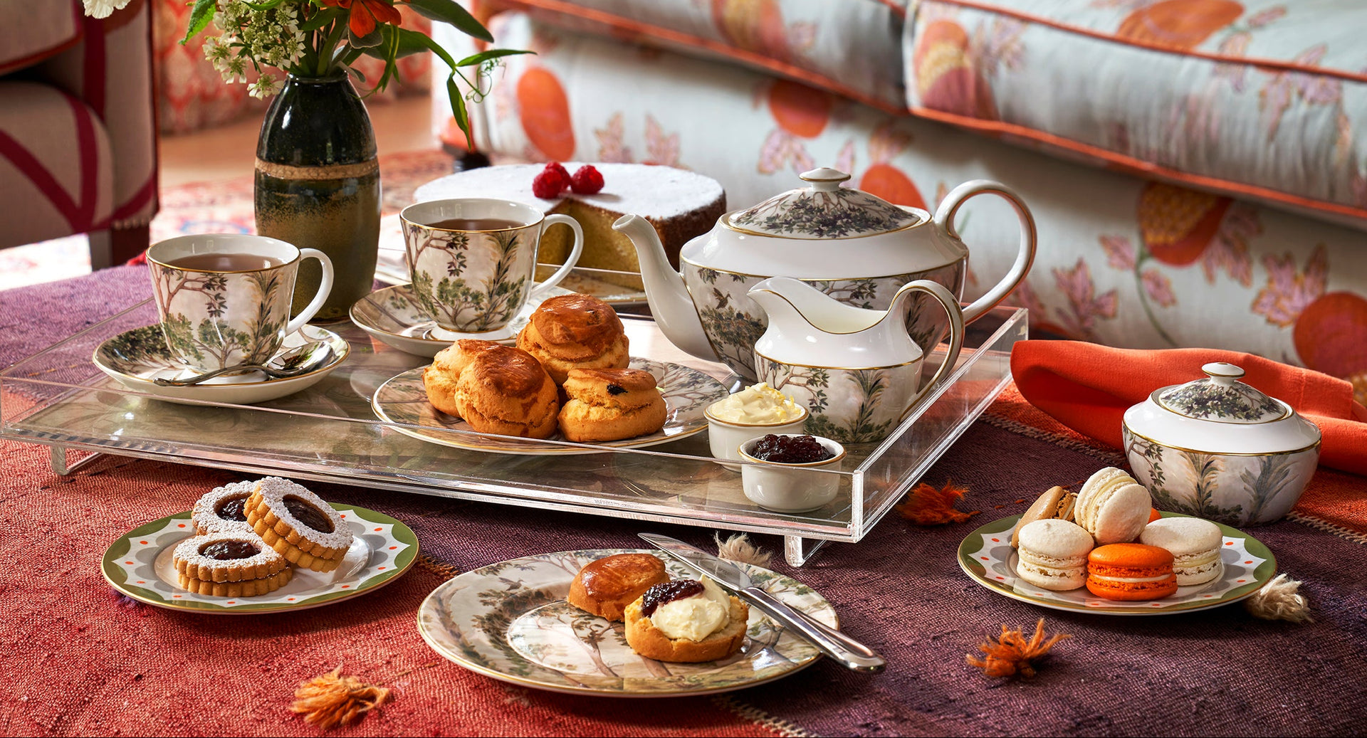Autumn afternoon tea collection by Shop Kit Kemp showcasing Tall Trees fine bone china teacups, teapot, creamer, and sugar bowl, all arranged on a signature acrylic tray inlaid with Kit Kemp fabric, presented on a table with other treats and autumnal decorations.