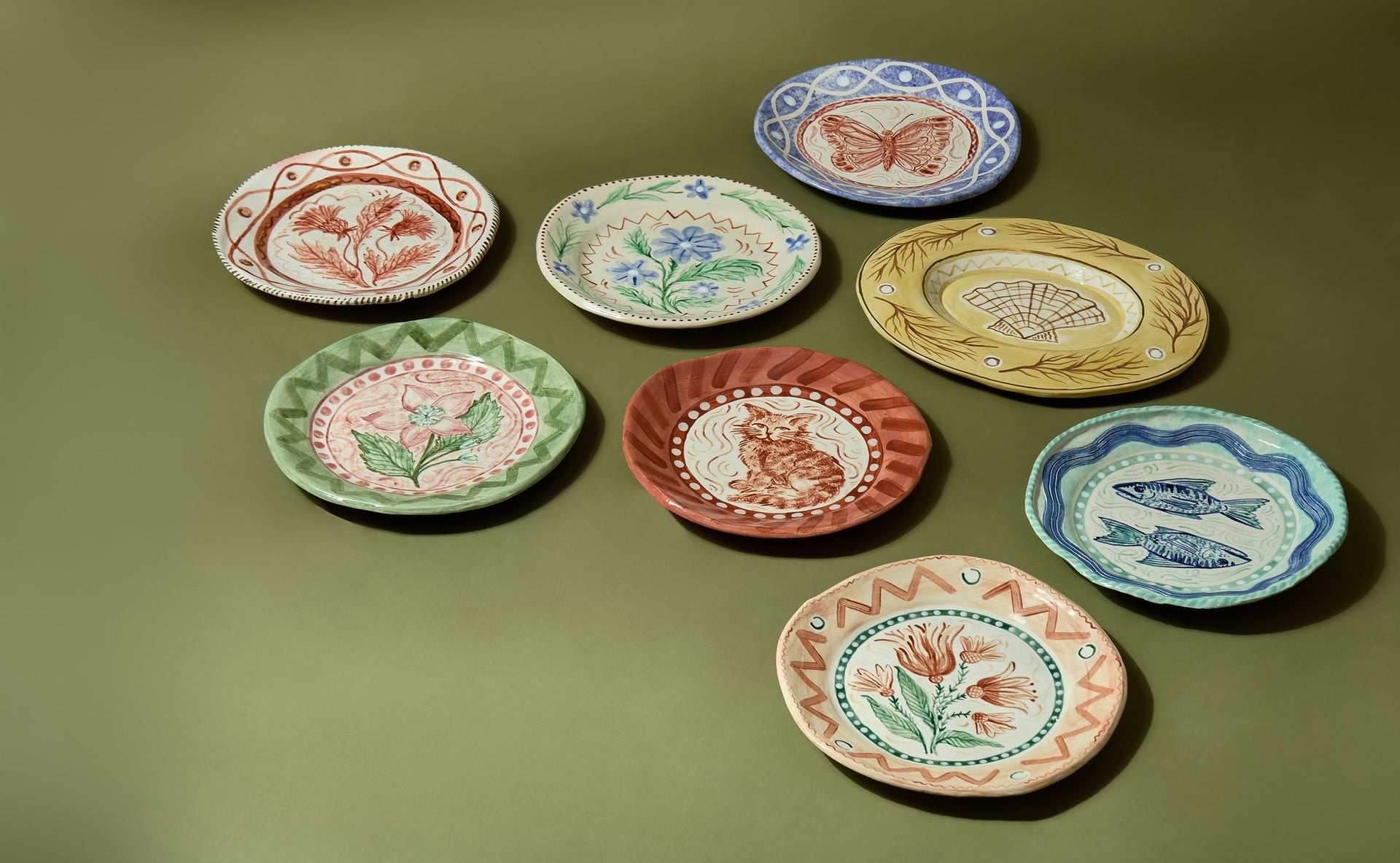 A curated lifestyle arrangement of hand-painted ceramic plates and bowls by artist Sasha Compton for Shop Kit Kemp, displayed against a sage green background. The collection features whimsical illustrations of a marmalade cat, blue butterfly, pink hellebores, and blue fish, all tied together by vibrant, freehand borders and scalloped edges.