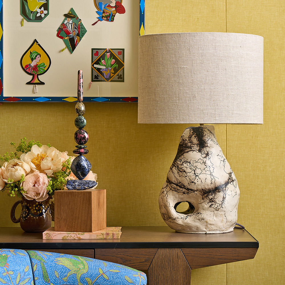 An organic-shaped, hand-sculpted ceramic table lamp base by artist Daniel Clarke for Shop Kit Kemp. The lamp features a raku-fired finish, and it is paired with a neutral drum linen lampshade and styled on a Kit Kemp desk.