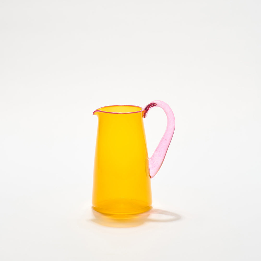A vibrant hand-blown glass jug with a transparent orange body and a pink handle, with a pale pink lip, by Shop Kit Kemp.