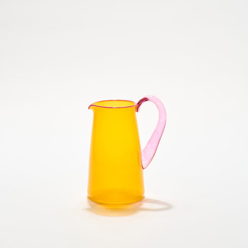A vibrant hand-blown glass jug with a transparent orange body and a pink handle, with a pale pink lip, by Shop Kit Kemp.