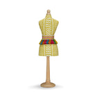 A handmade paper-mâché mini mannequin covered in a textured yellow "Tweak" fabric with a chevron-like weave. It is cinched at the waist with a fun, multicolour tassel belt and stands on a light wood base.
