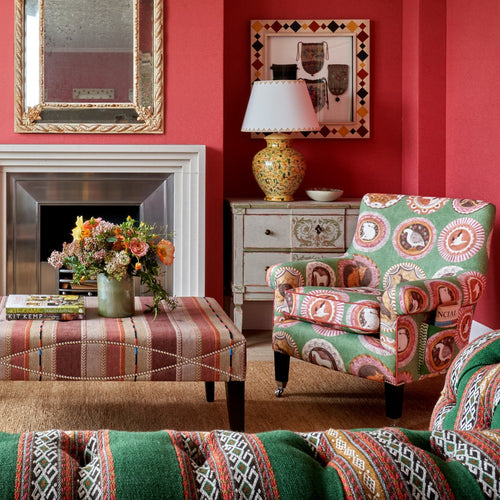 A pair of green Robina's Dinner Party Leo chairs are shown here displayed around a fireplace in front of Kit Kemp's Celia Ottoman in a wide striped brown cloth, with embroidery details, in a red room.