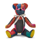 The image shows a stuffed teddy bear made from colourful patchwork fabric. The bear has a predominantly red, blue, and yellow colour scheme, with the fabric pieces arranged in a geometric pattern across its body. It is wearing a decorative ribbon around its neck. The bear is sitting upright, with its arms and legs splayed out to the sides, giving it a playful and inviting appearance. 