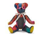 The image shows a stuffed teddy bear made from colourful patchwork fabric. The bear has a predominantly red, blue, and yellow colour scheme, with the fabric pieces arranged in a geometric pattern across its body. It is wearing a decorative ribbon around its neck. The bear is sitting upright, with its arms and legs splayed out to the sides, giving it a playful and inviting appearance. 