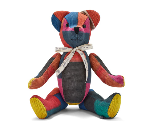 The image shows a stuffed teddy bear made from colourful patchwork fabric. The bear has a predominantly red, blue, and yellow colour scheme, with the fabric pieces arranged in a geometric pattern across its body. It is wearing a decorative ribbon around its neck. The bear is sitting upright, with its arms and legs splayed out to the sides, giving it a playful and inviting appearance. 