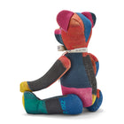 The image shows the back of a stuffed teddy bear made from colourful patchwork fabric. The bear has a predominantly red, blue, and yellow colour scheme, with the fabric pieces arranged in a geometric pattern across its body. It is wearing a decorative ribbon around its neck. The bear is sitting upright, with its arms and legs splayed out to the sides, giving it a playful and inviting appearance. 