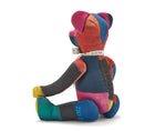 The image shows the back of a stuffed teddy bear made from colourful patchwork fabric. The bear has a predominantly red, blue, and yellow colour scheme, with the fabric pieces arranged in a geometric pattern across its body. It is wearing a decorative ribbon around its neck. The bear is sitting upright, with its arms and legs splayed out to the sides, giving it a playful and inviting appearance. 