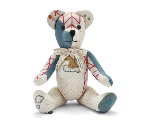 The image shows a stuffed teddy bear designed with various colourful patchwork fabrics. The bear has a mix of patterns and colours, including pink and orange stripes on the face and ears, solid blue on the left arm, and orange polka dots on other parts of the body. There are also stars on one ear and clouds on one foot. A ribbon is tied around its neck. There is a small picture of a dog with wings sitting on a cloud on its chest.