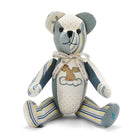 The image shows a stuffed teddy bear designed with various colourful patchwork fabrics. The bear has a mix of patterns and colours, including blue, yellow and white stripes on the face, legs and hands and orange polka dots on one arm and tummy. There are also stars on one ear. A ribbon is tied around its neck. There is a small picture of a dog with wings sitting on a cloud on its chest.