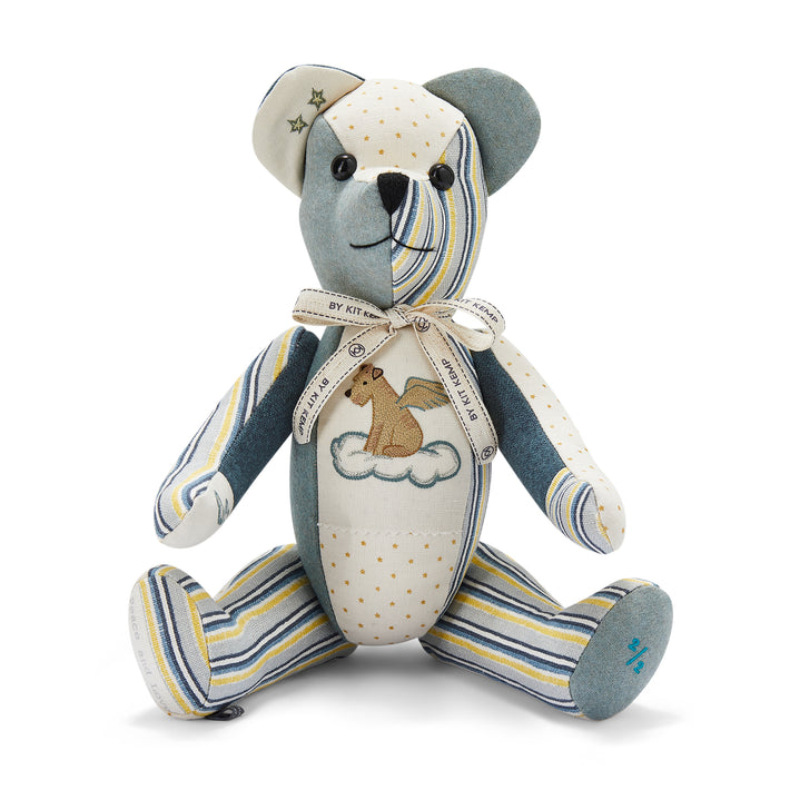 The image shows a stuffed teddy bear designed with various colourful patchwork fabrics. The bear has a mix of patterns and colours, including blue, yellow and white stripes on the face, legs and hands and orange polka dots on one arm and tummy. There are also stars on one ear. A ribbon is tied around its neck. There is a small picture of a dog with wings sitting on a cloud on its chest.