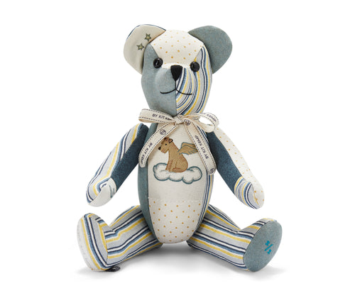 The image shows a stuffed teddy bear designed with various colourful patchwork fabrics. The bear has a mix of patterns and colours, including blue, yellow and white stripes on the face, legs and hands and orange polka dots on one arm and tummy. There are also stars on one ear. A ribbon is tied around its neck. There is a small picture of a dog with wings sitting on a cloud on its chest.