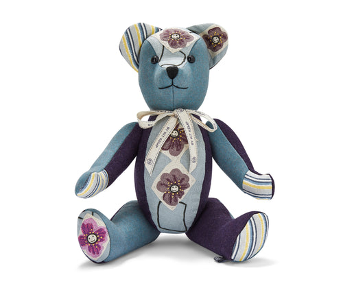 The image shows a stuffed teddy bear toy. The bear has blue fabric with floral patterns on its body. The bear's arms and legs are made of a darker blue fabric with yellow, blue and white stripes on its hands and one foot. Around its neck is a ribbon tied in a bow. The bear is sitting upright and handmade from various fabric pieces sewn together to create a charming, decorative stuffed animal.