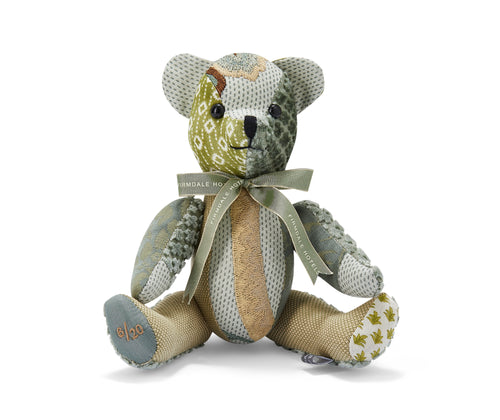 The image depicts a plush toy in the shape of a teddy bear. The bear is made from a patchwork of different fabrics, including a patterned green and white fabric, and a tan-coloured material. The bear is wearing a green ribbon around its neck, which has the text Firmdale Hotels on it. 