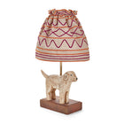 The Kit Kemp Doggy lamp is a small table lamp, consisting of a naively sculpted dog standing on the dark rectangular base of lamp, under a ruched natural linen shade with burgundy and orange stitch work forming horizontal stripes and looping patterns on the gathered shade.