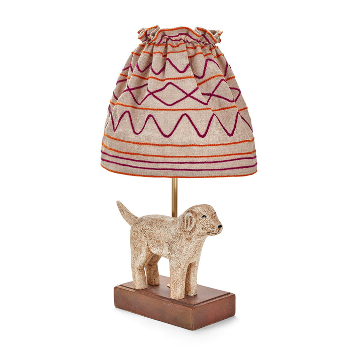 The Kit Kemp Doggy lamp is a small table lamp, consisting of a naively sculpted dog standing on the dark rectangular base of lamp, under a ruched natural linen shade with burgundy and orange stitch work forming horizontal stripes and looping patterns on the gathered shade.