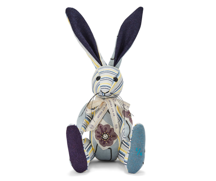 The image shows a stuffed toy rabbit. The rabbit has blue fabric with floral patterns on its body. The rabbit has long ears that are navy in colour. Its face and limbs are partly covered in a striped fabric with yellow, blue and white stripes. Around its neck is a ribbon tied in a bow. The rabbit is sitting upright and handmade from various fabric pieces sewn together to create a charming, decorative stuffed animal. 