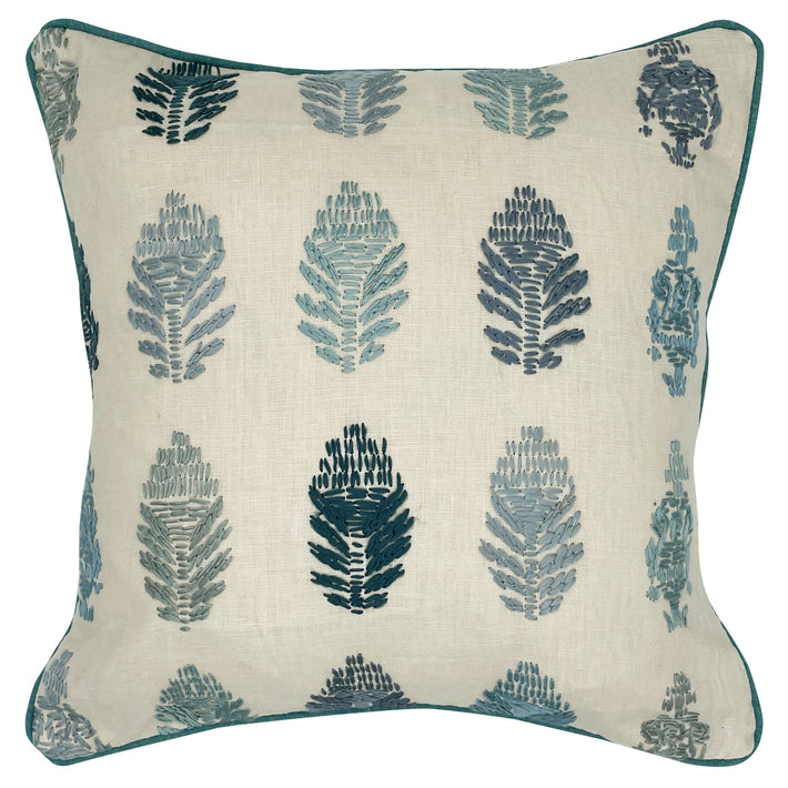 The image displays a cushion with a botanical design. The pattern features stylized stitched representations of fern-like plants in two colors, dark blue and a lighter blue grey, set against a neutral beige fabric background. 