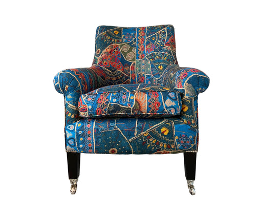 The Polly armchair is covered in Kit kemps new fabric ,with Gp & J Baker, in blue, with its patchwork of intricate patterns in reds, yellows , and shades of blue.. shown from the front, with its dark wooden legs and brass wheel feet.