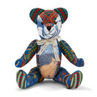 The image shows a colourful, patchwork-style teddy bear. The bear is designed with various fabric patterns, including a cat motif on its tummy. It has a ribbon around its neck that reads BY KIT KEMP. The teddy bear's body features vibrant, multicoloured designs, giving it a distinct and artistic appearance.