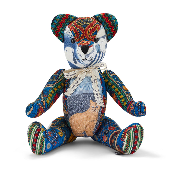 The image shows a colourful, patchwork-style teddy bear. The bear is designed with various fabric patterns, including a cat motif on its tummy. It has a ribbon around its neck that reads BY KIT KEMP. The teddy bear's body features vibrant, multicoloured designs, giving it a distinct and artistic appearance.