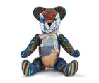 The image shows a colourful, patchwork-style teddy bear. The bear is designed with various fabric patterns, including a cat motif on its tummy. It has a ribbon around its neck that reads BY KIT KEMP. The teddy bear's body features vibrant, multicoloured designs, giving it a distinct and artistic appearance.