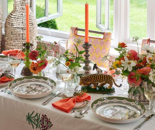 The Tall Tree and Geo large pasta bowl is shown on a table dressed for lunch with bright orange napkins, and a floral embroidered white tablecloth. The table is also dressed with candlesticks by Katrin Moye.