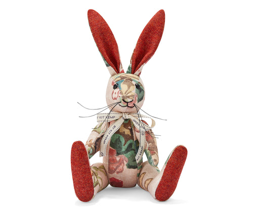 The image shows a stuffed toy rabbit with long ears. The rabbit has a floral pattern fabric covering its body, featuring red, green and cream coloured flowers and leaves. It is wearing a ribbon tied in a bow around its neck, with text printed on the ribbon that says By Kit Kemp. The rabbit is sitting upright. Its long ears and feet are a solid reddish colour that contrasts with the floral patterned body.