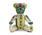 The image shows a stuffed teddy bear made from patterned fabric. The bear has a primarily white body with green, blue, red and yellow geometric patterns and designs covering it. 3D rings are stitched down the front of the bears tummy. A ribbon is tied around the bear's neck that says BY KIT KEMP. The patterns on the fabric resemble abstract leaves and vines in an earthy colour palette. The bear's limbs are posed in a sitting position