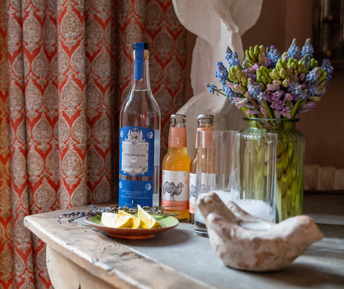 Life style shot of our bespoke gin gift set, presented with the 2 glasses and grapefruit tonic water, contained in the gift box, presented with ice, and lemon, surrounded by spring flowers in a vase.